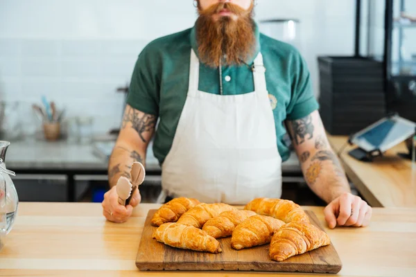 Hombre barbudo en delantal de pie y sosteniendo pinzas para croissants —  Fotos de Stock