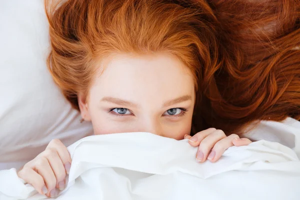 Jolie femme mignonne aux cheveux roux se cachant sous une couverture blanche — Photo