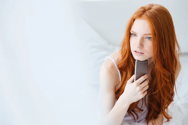 Thoughtful woman holding mobile phone and thinking in bed — Stock Photo, Image