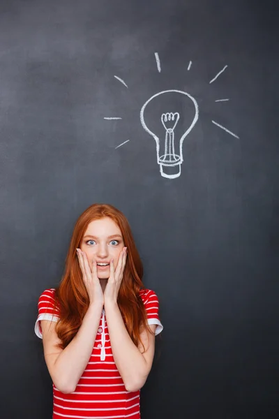 Amazed woman standing over chalkboard background with drawn electric bulb — 스톡 사진