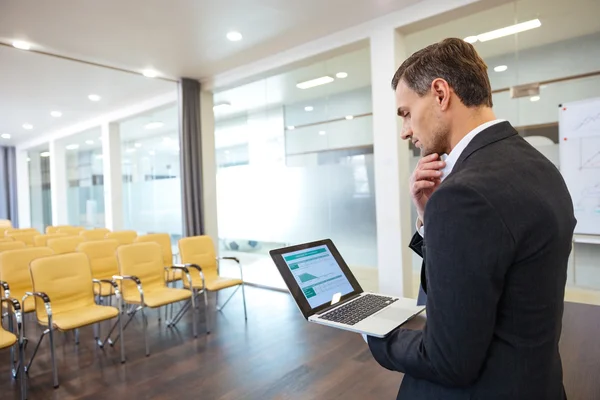 Fundersam affärsman med laptop står i tomma konferenslokal — Stockfoto