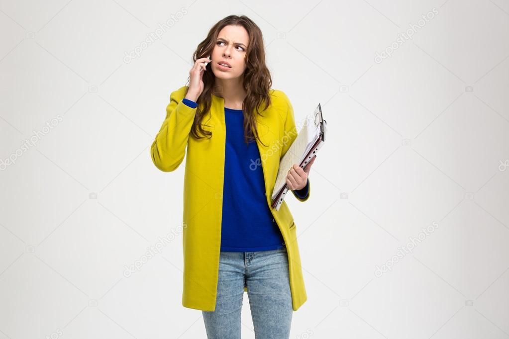 Thoughtful frowning young woman holding clipboard and using smartphone