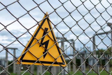 Metal ağ çitlerinin arkasında yıldırımlı sarı üçgen, yüksek voltajlı uyarı işareti