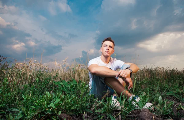 Zamyślony przystojny młody mężczyzna siedzący na zielonej trawie na ba — Zdjęcie stockowe
