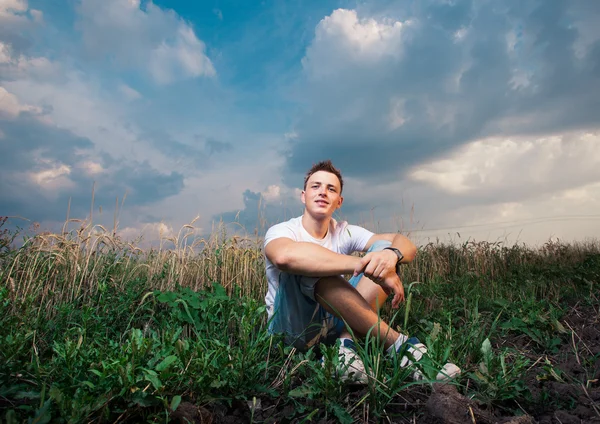 Uśmiechający się przystojny młody mężczyzna siedzący na zielonej trawie na ba — Zdjęcie stockowe