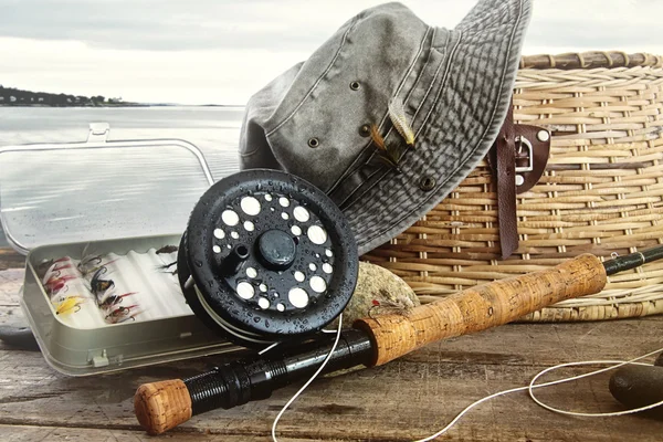 Sombrero y artes de pesca con mosca en la mesa cerca del agua — Foto de Stock