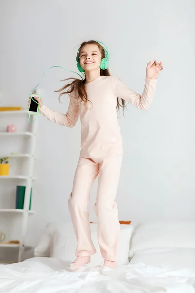 Chica saltando en la cama con teléfono inteligente y auriculares —  Fotos de Stock