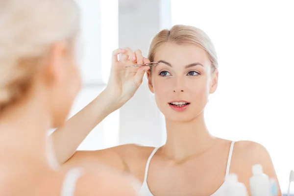 Woman with tweezers tweezing eyebrow at bathroom — Stock Photo, Image