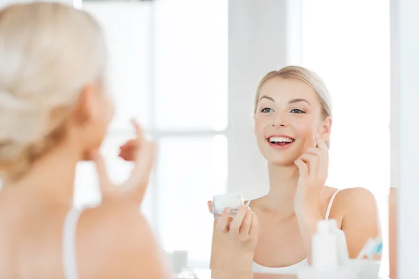 Happy woman applying cream to face at bathroom — Stock Photo, Image