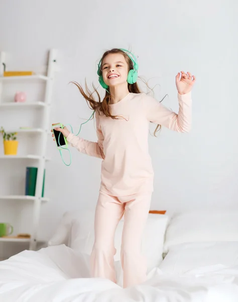 Chica saltando en la cama con teléfono inteligente y auriculares —  Fotos de Stock