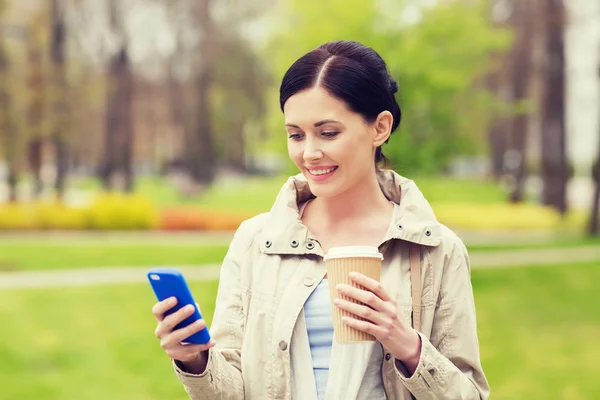 Mujer sonriente con teléfono inteligente y café en el parque — Foto de Stock