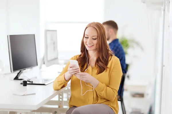 Kvinne med øretelefoner og smarttelefon ved kontoret – stockfoto