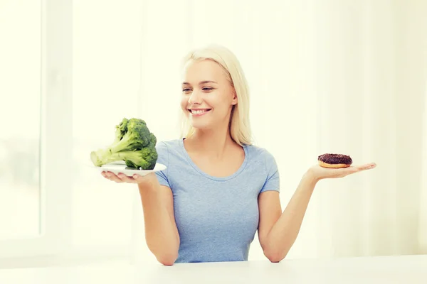 Leende kvinna med broccoli och donut hemma — Stockfoto