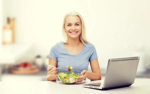 Donna sorridente mangiare insalata con computer portatile in cucina — Foto Stock