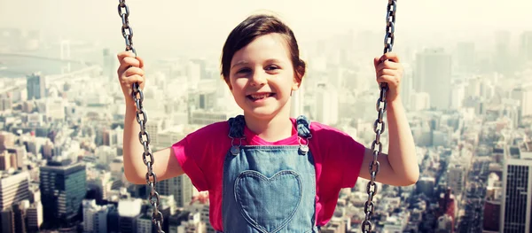 Happy little girl swinging on swing over city — Stockfoto