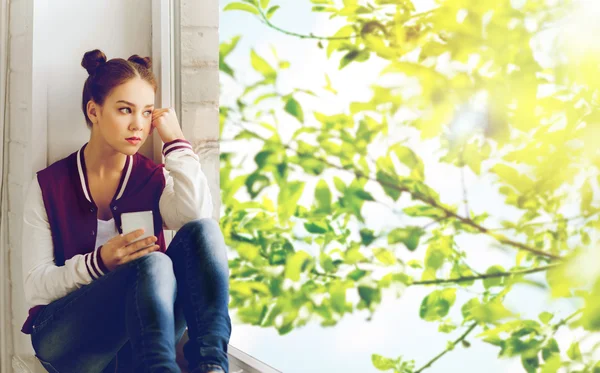 Chica adolescente sentada en el alféizar de la ventana con teléfono inteligente — Foto de Stock