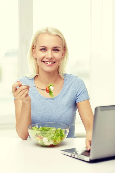 Donna sorridente con computer portatile mangiare insalata a casa — Foto Stock