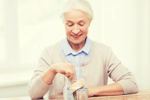 Donna anziana mettendo soldi in vaso di vetro a casa — Foto Stock