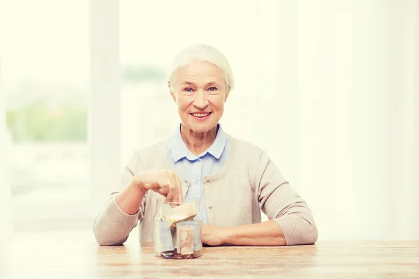 senior woman putting money into glass jar at home