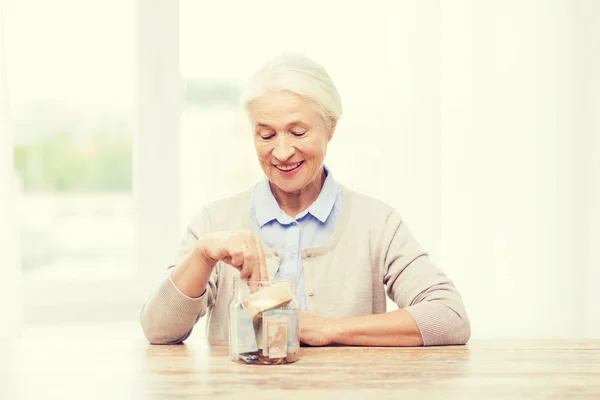 Старша жінка кладе гроші в скляну банку вдома — стокове фото