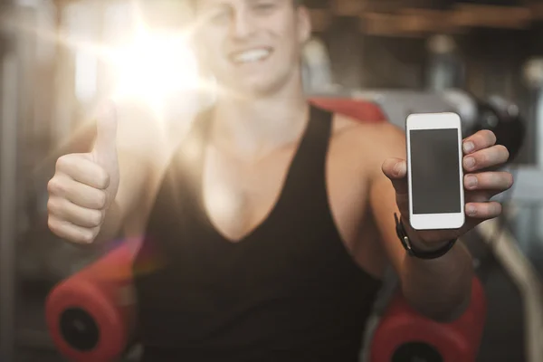 Junger Mann mit Smartphone zeigt im Fitnessstudio Daumen hoch — Stockfoto