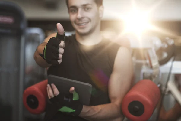 Young man with tablet pc showing thumbs up in gym — Stock Photo, Image