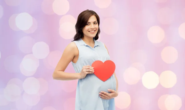 Happy těhotná žena s červeným srdcem dotýká břicho — Stock fotografie