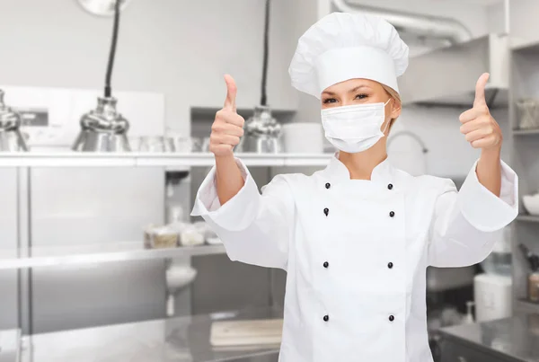 Kobieta szef kuchni w masce pokazując kciuki w górę w kuchni — Zdjęcie stockowe