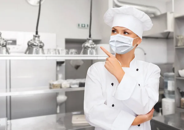 Mujer chef en máscara apuntando dedo hacia arriba en la cocina —  Fotos de Stock