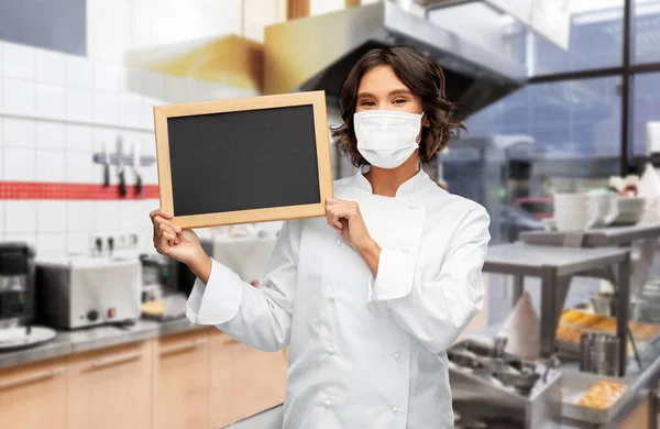 Női szakács maszkban táblával a konyhában — Stock Fotó