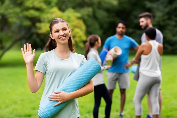 Mujer sonriente con esterilla de yoga agitando la mano en el parque — Foto de Stock