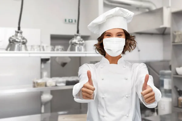 Női szakács maszkban mutatja hüvelykujját fel a konyhában — Stock Fotó