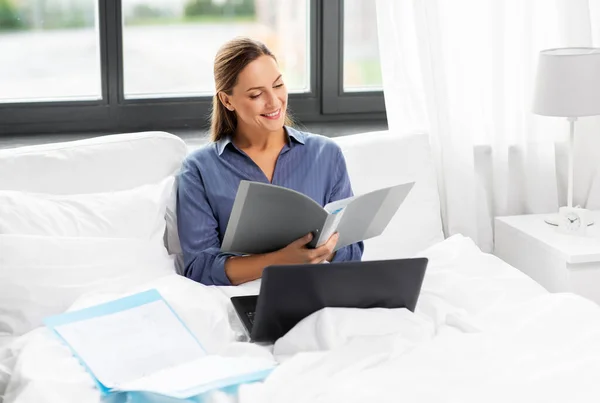 Mujer joven con portátil y papeles en la cama en casa —  Fotos de Stock
