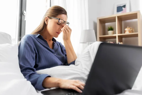 Mujer cansada en gafas con portátil en la cama en casa —  Fotos de Stock