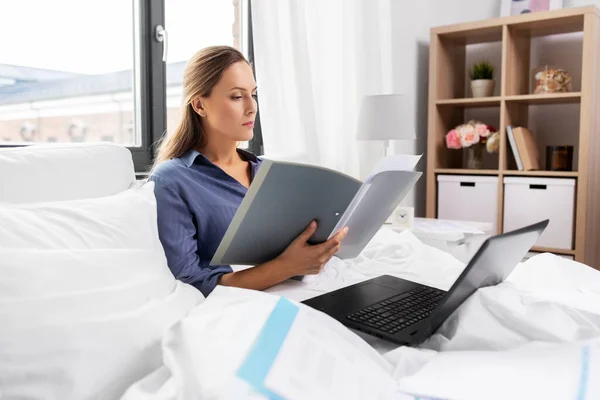 Jonge vrouw met laptop en papieren in bed thuis — Stockfoto