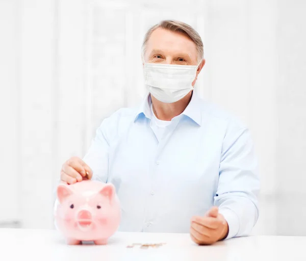 Velho homem em máscara colocando moeda em grande banco porquinho — Fotografia de Stock