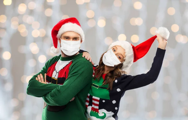 Casal em máscaras, camisolas de Natal e chapéus de santa — Fotografia de Stock