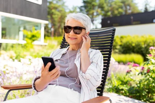 Starsza kobieta ze słuchawkami i smartfonem w ogrodzie — Zdjęcie stockowe