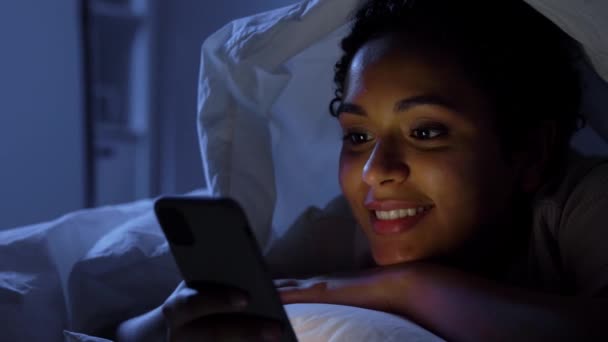 Жінка зі смартфоном під ковдрою в ліжку вночі — стокове відео