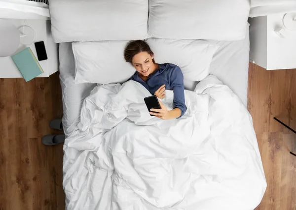 Молодая женщина со смартфоном лежит в постели — стоковое фото