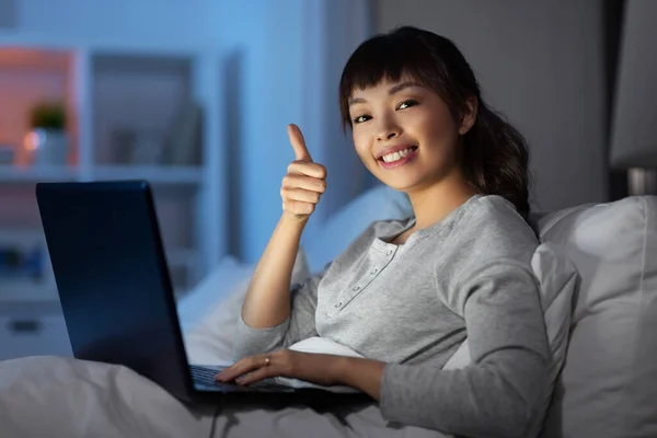 Mulher com laptop na cama à noite mostra polegares para cima — Fotografia de Stock