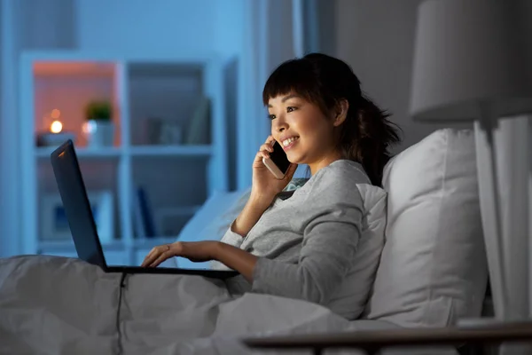 Mujer con portátil llamando por teléfono en la cama por la noche — Foto de Stock