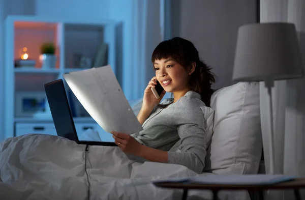 Mujer con portátil llamando por teléfono en la cama por la noche —  Fotos de Stock