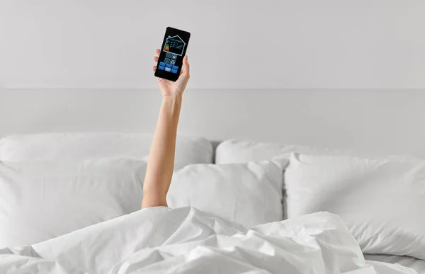 Mano en la cama con iconos de casa inteligente en el teléfono inteligente —  Fotos de Stock