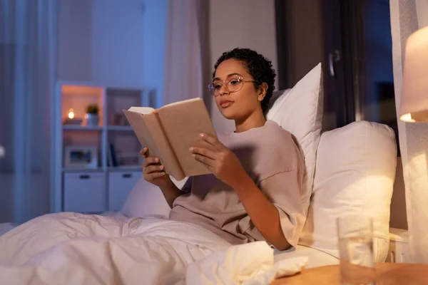 Mujer joven en gafas lectura libro en la cama en casa —  Fotos de Stock