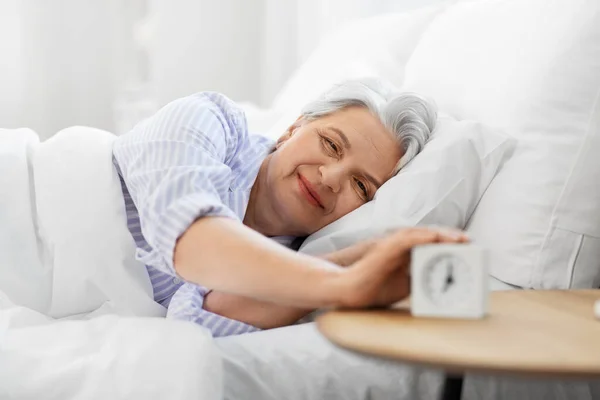 Счастливая пожилая женщина с будильником в постели дома — стоковое фото