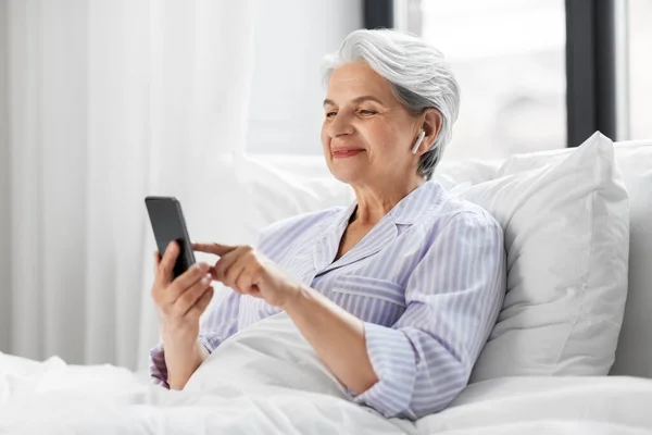 Donna anziana con smartphone e auricolari a letto — Foto Stock