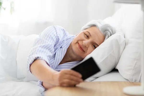 Счастливая пожилая женщина со смартфоном в постели дома — стоковое фото