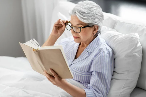 Old woman in glasses reading book in bed at home — Stock Photo, Image