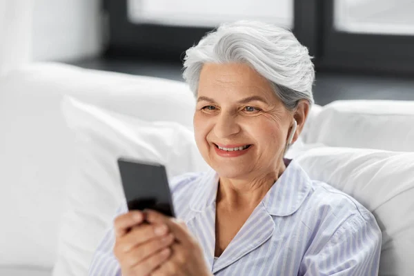 Mulher idosa com smartphone e fones de ouvido na cama — Fotografia de Stock